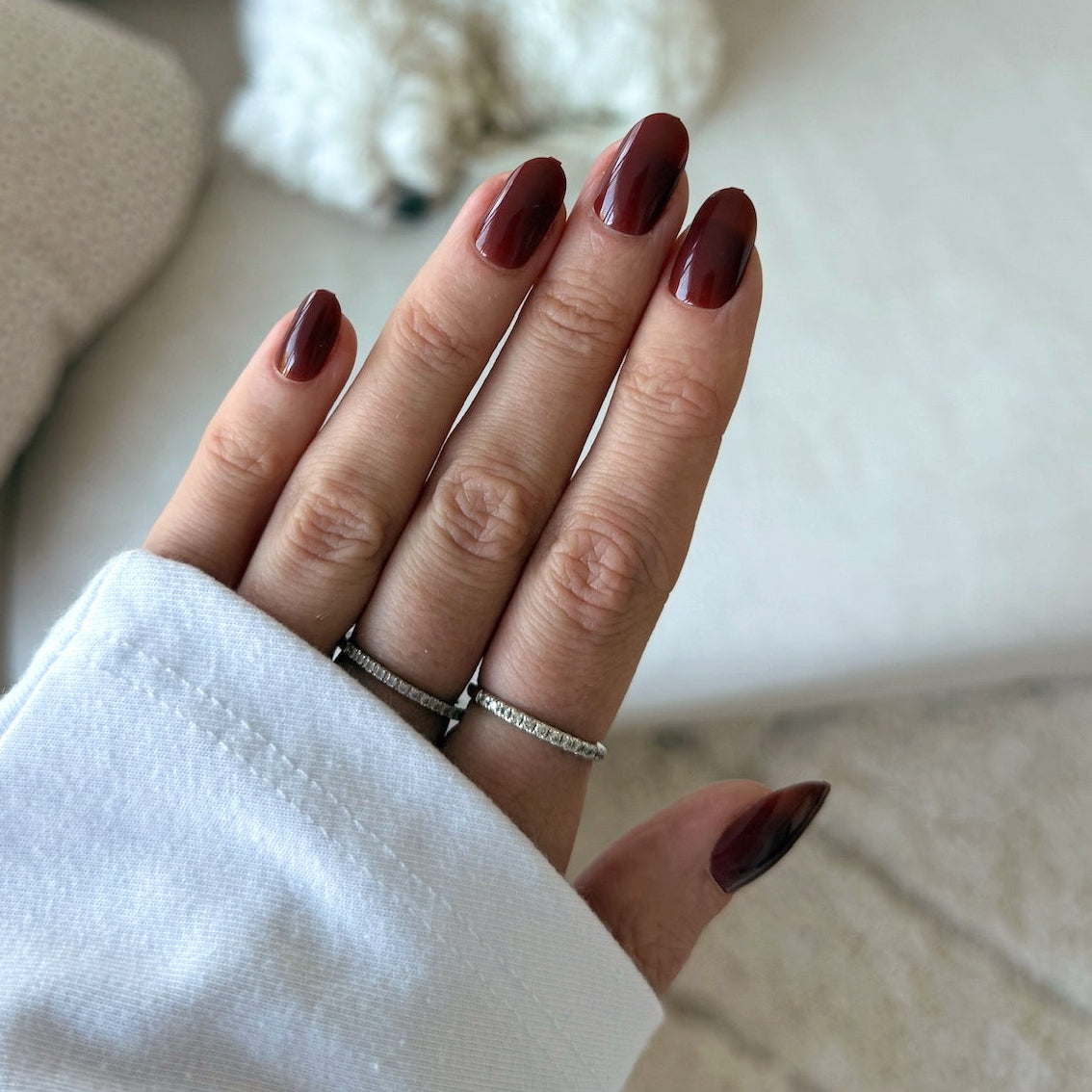 Close-up of a hand with maroon nail polish wearing two rings against a blurred indoor background.