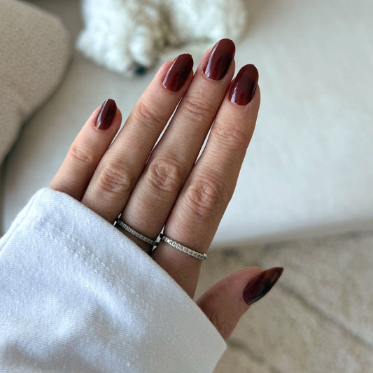 Close-up of a hand with maroon nail polish wearing two rings against a blurred indoor background.